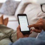 Closeup of a man hand holding cellphone with internet browser on screen. Man with spectacles relaxing sitting on couch while looking at mobile phone. Closeup of mature latin man using smartphone to checking email at home. (Closeup of a man hand holdin